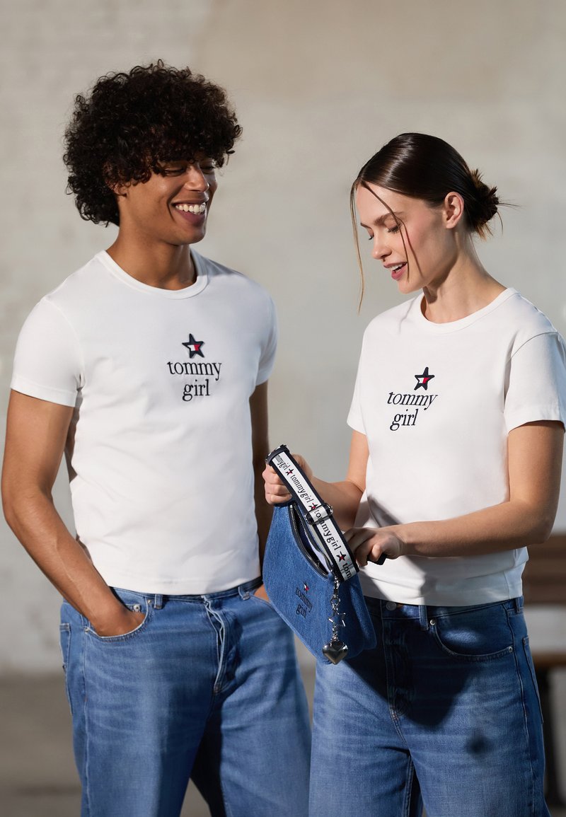 Hvite t-skjorter med "tommy girl"-logo og stjerne. Lys blå denimjeans. Kvinnen holder en blå denimveske med en svart stropp og hjerteanheng.