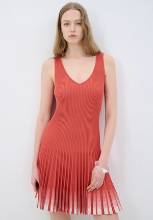 Ribbed sleeveless dress in coral with a V-neckline, featuring a pleated skirt that fades to white at the hem. Smooth texture, minimalist design.