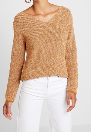 Femme portant un pull texturé orange à col en V associé à un pantalon blanc taille haute et de grandes boucles d'oreilles.