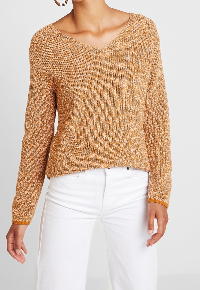 Femme portant un pull texturé orange à col en V associé à un pantalon blanc taille haute et de grandes boucles d'oreilles.