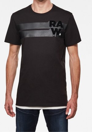 Mann trägt ein schwarzes kurzärmliges T-Shirt mit zwei horizontalen dunkelgrauen Streifen und dem auffälligen Schriftzug "RAW." auf der oberen linken Brust.