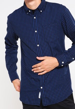 Mężczyzna w niebiesko-czarnej koszuli w kratę zapinanej na guziki, jedną ręką na brzuchu, drugą przy boku.