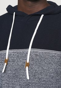 Marineblauwe hoodie met een gestructureerd grijs onderpaneel, witte trekkoorden en bruine plastic accenten. Bevat een geribbelde ontwerpelementen en contrasterende strepen.
