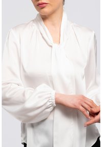 Weißes Bluse aus weichem, leichtem Stoff. Eigenschaften: gebundener Ausschnitt und voluminöse Ballonärmel mit elastischen Bündchen.