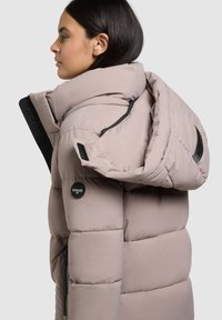 Puffige, hellbeige Jacke mit hohem Kragen und großer Kapuze. Verfügt über schwarze Reißverschlusstaschen und ein Logo-Patch am Ärmel. Glatte Textur.