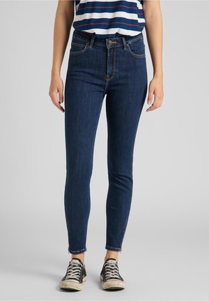 Los jeans de mezclilla azul ajustados cuentan con una cintura alta, un diseño clásico de cinco bolsillos y cremalleras en los tobillos. Se combinan con zapatillas negras y una blusa a rayas.