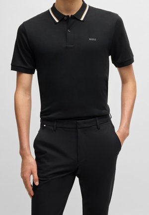 Mann trägt schwarzes Poloshirt mit kurzen Ärmeln, gestreiftem Kragen und „BOSS“-Logo, kombiniert mit schwarzen Hosen, Hände in den Taschen.