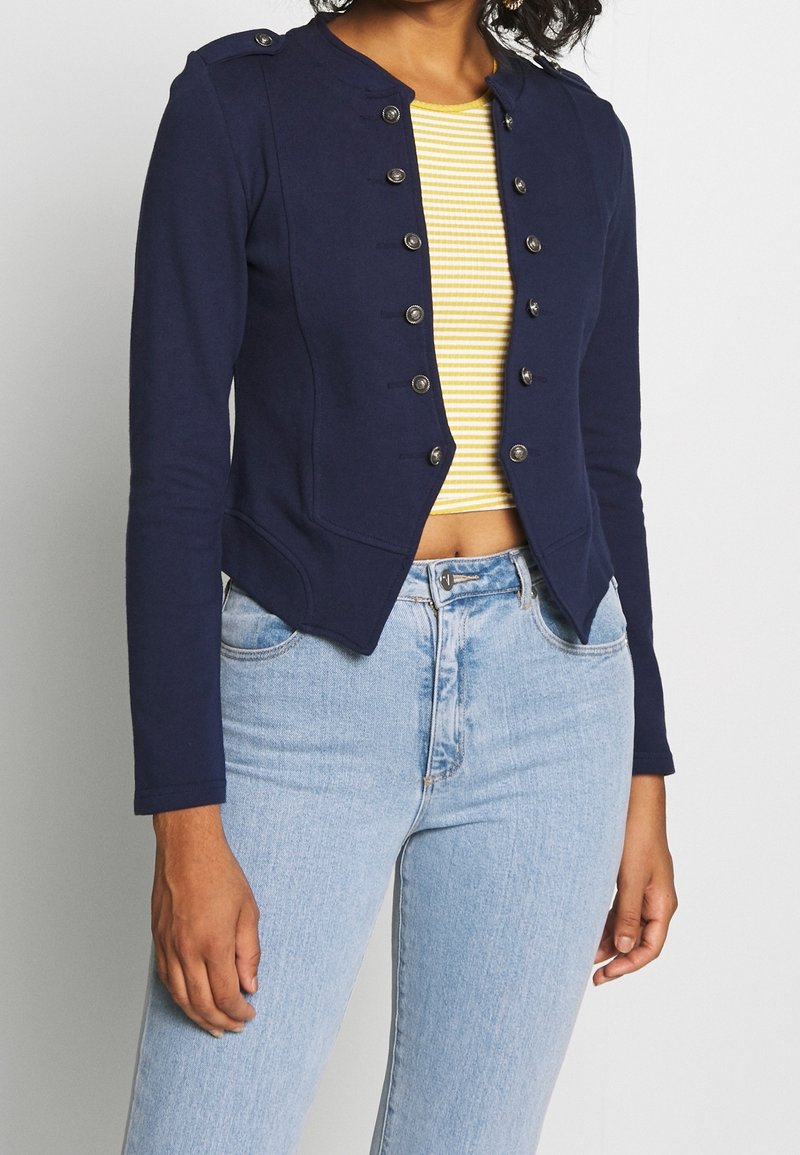 Femme portant une veste militaire bleu marine avec des boutons argentés, par-dessus un crop top jaune à rayures et un jean taille haute bleu clair.