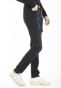 Jean cargo noirs avec une coupe slim, dotés de poches latérales et d'un design de jambe légèrement effilée. Portés avec des baskets blanches sur un fond uni.