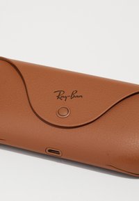 Custodia per occhiali da sole in pelle marrone con una texture liscia, dotata di una chiusura a pattina curva e un logo impresso. Include una porta di ricarica USB-C.