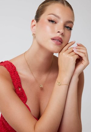 Set di gioielli in oro composto da una collana e un bracciale con pendenti circolari, abbinati a orecchini a stud. Il modello indossa un top rosso all'uncinetto.