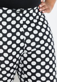 Zwarte stoffen broek met een patroon van grote witte stippen, met een knoopsluiting en zijzakken. Ee smooth textuur.