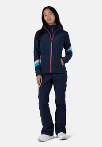 Giacca in pile blu navy con colletto alto, zip rosa e strisce colorate sulle maniche, abbinata a pantaloni blu navy e calzature bianche.