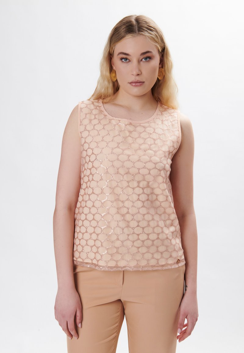Top senza maniche color pesco con un motivo a rete esagonale, texture liscia e una leggera lucentezza, abbinato a pantaloni aderenti di colore chiaro.