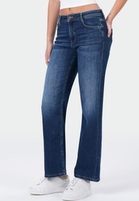 Vrouw in mid-rise recht gesneden donkerblauwe jeans en witte sneakers, staand met één hand in haar zak tegen een effen achtergrond.