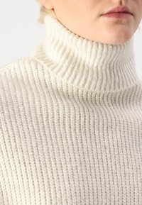 Kremasta turtleneck pulover z rebrastim vzorcem, ki ima visok ovratnik in tesno prileganje.