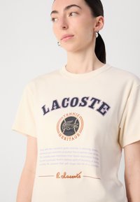 Bež majica od pamuka s okruglim izrezom, kratkim rukavima i ispisanim logotipom "LACOSTE" s grafičkim naglasima u mornarsko plavoj i narančastoj boji.
