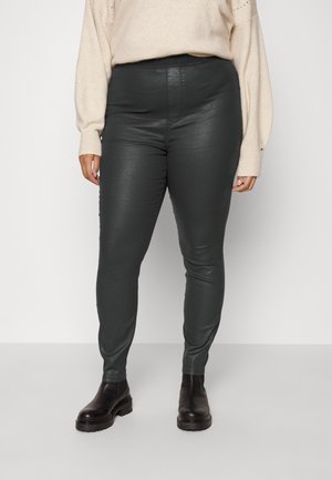 Personne portant un pantalon noir ajusté taille haute, un pull beige à manches longues et des bottines noires, debout sur un fond uni.