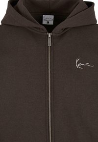 Sudadera con cremallera de color marrón oscuro hecha de algodón suave, con capucha forrada y un logo bordado en blanco en el pecho, detalle de cremallera plateada.