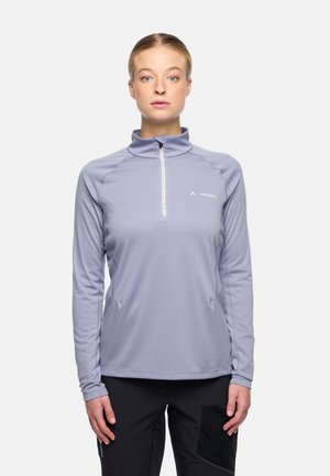 Femme portant un haut de sport violet clair à manches longues avec fermeture éclair et un pantalon noir, se tenant devant un fond neutre.