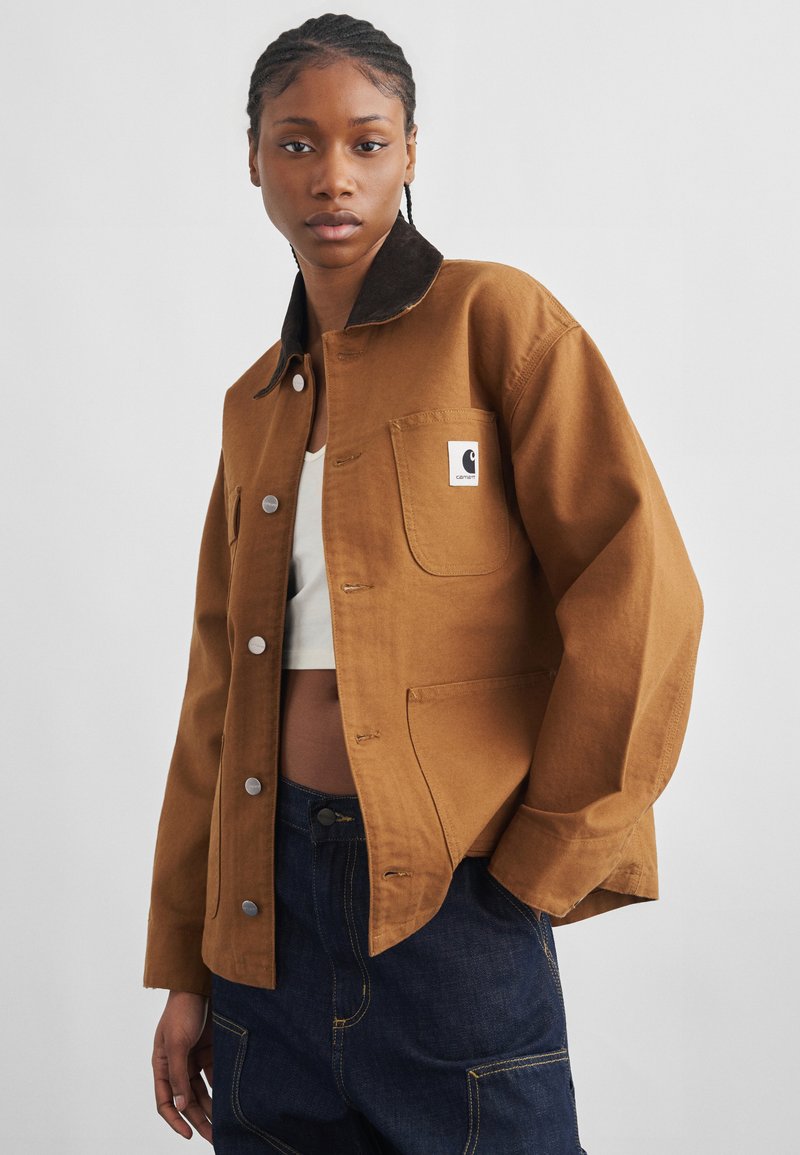 Giovane con capelli intrecciati che indossa una giacca marrone con bottoni sopra un top corto bianco e jeans blu scuro davanti a uno sfondo semplice.