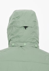 Ljusgrön huva jacka med en texturerad yta, som har en dragsko bak och tryckknappar vid halsen. Enkel, funktionell design.