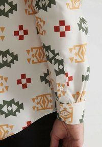 Chemise blanche avec des motifs géométriques en vert, rouge et orange, comportant un poignet à boutons et une texture de tissu lisse.