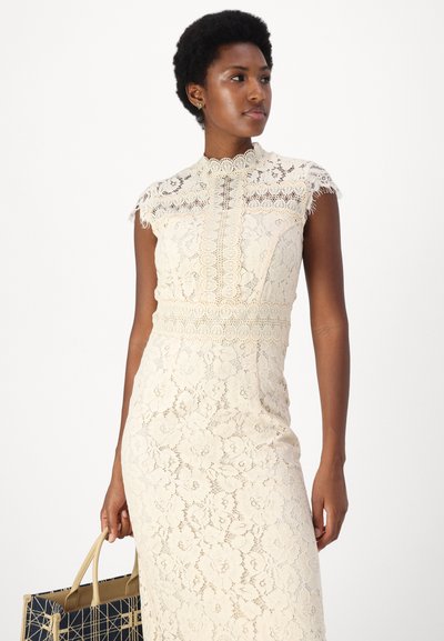 Robe en dentelle crème avec un col haut et des manches courtes, ornée de motifs floraux et de bords festonnés, accompagnée d'un sac à main à motifs.