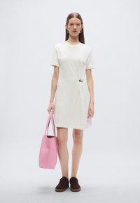 Λευκό βαμβακερό φόρεμα τύπου T-shirt με κοντά μανίκια, διακοσμημένο με πράσινη λεπτομέρεια. Το μοντέλο κρατά ροζ τσάντα tote και φορά καφέ παπούτσια. Απλός σχεδιασμός.