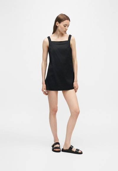 Jeune femme aux cheveux bruns lisses portant une mini-robe noire sans manches et des sandales noires, debout devant un fond blanc uni.