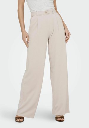 Femme portant un pantalon beige taille haute à jambes larges avec une ceinture assortie et des sandales ouvertes marron, debout devant un fond blanc.