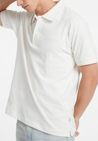 Polo shirt bianco in cotone morbido con colletto classico e maniche corte. Presenta il logo discreto di Guess Jeans, spacco sui fianchi e vestibilità rilassata.