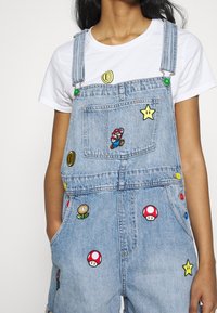 Salopette en jean avec des personnages et symboles brodés de Super Mario, portée sur un t-shirt blanc uni.