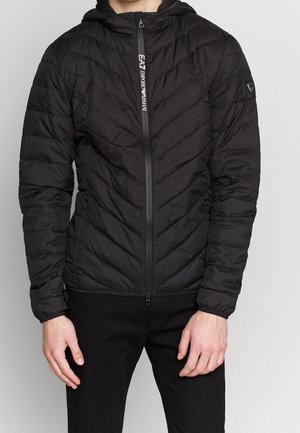 Mężczyzna ubrany w czarną pikowaną kurtkę zapinaną na zamek z kapturem i logo „EA7 Emporio Armani” w okolicy zamka, w połączeniu z czarnymi spodniami.