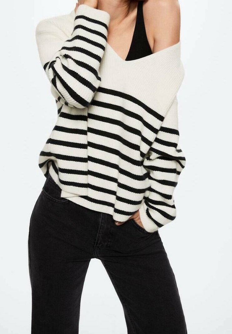 Femme portant un pull blanc à épaules dénudées avec des rayures horizontales noires sur un haut noir, associée à un pantalon noir, posant avec une main dans la poche.