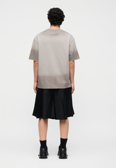 Άτομο με σγουρά μαλλιά στέκεται με την πλάτη, φοράει γκρι φαρδιά μπλούζα, μαύρη πλισέ φούστα μέχρι το γόνατο, μαύρες κάλτσες και μαύρα αθλητικά παπούτσια.