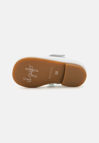 Zapato blanco con una forma suave y redondeada y una suela de goma color tostado. Presenta acentos metálicos y una marca en relieve en la suela.