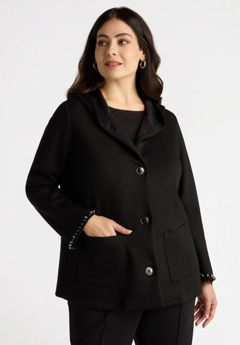 Chaqueta negra con cuello de chal, botones en el frente y dos bolsillos; presenta embellishments con tachuelas en los puños y una textura de tela suave.