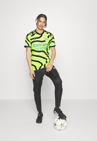 Camiseta de fútbol a rayas verdes y negras con acentos azules, combinada con pantalones negros. Zapatillas negras sobre un balón de fútbol blanco con estrellas de colores.