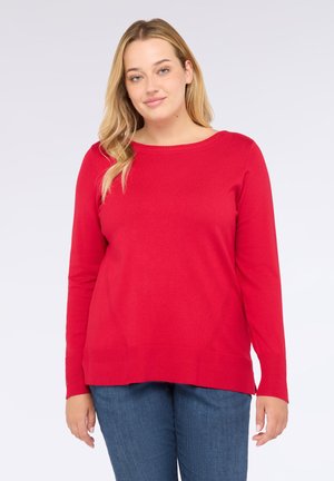 Mujer con cabello largo y rubio, vistiendo un suéter rojo de manga larga y jeans azules, de pie frente a un fondo claro y liso.