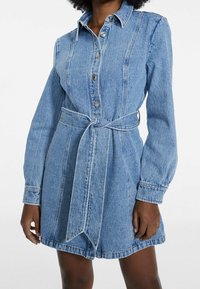 Denimmekko, jossa on kaulus, pitkät hihat ja nappikiinnitys. Vöissä on solmittava vyö, ja se on tyyliltään vaalean sininen pesu.