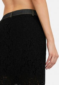 Jupe en dentelle noire avec un motif floral, dotée d'une taille en denim et de détails de couture. Le design texturé contraste avec le tissu lisse.