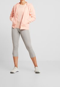Persikka vetoketjuhuppari sivutaskuilla, yhdistetty vaaleanharmaisiin capri-leggingseihin, joissa on sauman yksityiskohtia ja hienovarainen logo. Vaaleat tennarit.