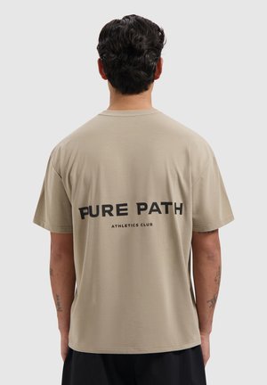 Man met donker haar die een beige T-shirt draagt met de tekst "PURE PATH ATHLETICS CLUB" op de achterkant, staand tegen een effen lichtgrijze achtergrond.