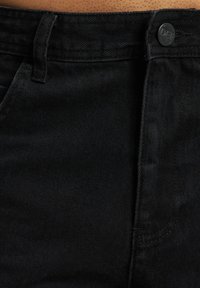 Jeans de mezclilla negra con cierre de botón, trabillas para cinturón y una textura suave. La costura a lo largo de los lados y los bolsillos es visible.