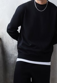Sudadera negra de algodón con bajo y puños acanalados, combinada con una camiseta blanca visible en la parte inferior, y un collar de cadena plateada.
