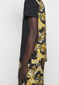 Person trägt ein schwarzes T-Shirt und dazu passende schwarze Shorts mit komplexen barocken Mustern in Gelb und Weiß sowie Text.