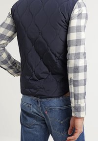 Chaleco acolchado en color navy con una textura suave, usado sobre una camisa de cuadros en gris y blanco. Detalles de costura visibles y un ajuste casual.