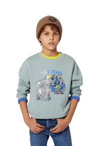 Sweatshirt teal claro com um gráfico de urso e uma cena de montanha, com punhos canelados em azul, sobre uma t-shirt amarela, combinado com jeans azuis.