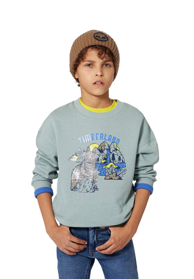 Sweatshirt teal claro com um gráfico de urso e uma cena de montanha, com punhos canelados em azul, sobre uma t-shirt amarela, combinado com jeans azuis.